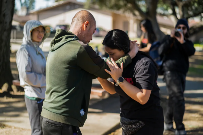 A light-skinned man with a shaven head, wearing a green hoodie, wraps his arms atop a woman's shoulders with his hands clasped behind her neck. The woman leans forward and holds onto both of the man's arms.