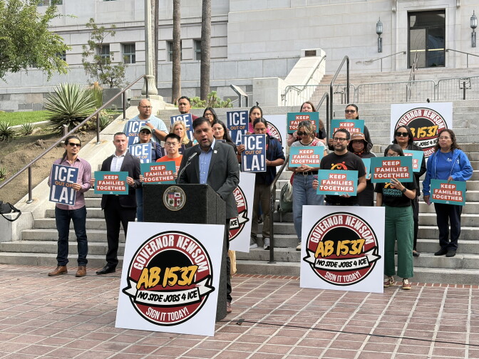 A man with short dark hair and medium light skin tone is dressed in a light blue collared shirt and sports coat, speaking at a podium. Behind him, about twenty people hold up signs that read "ICE out of L.A." and "Keep Families Together." They 