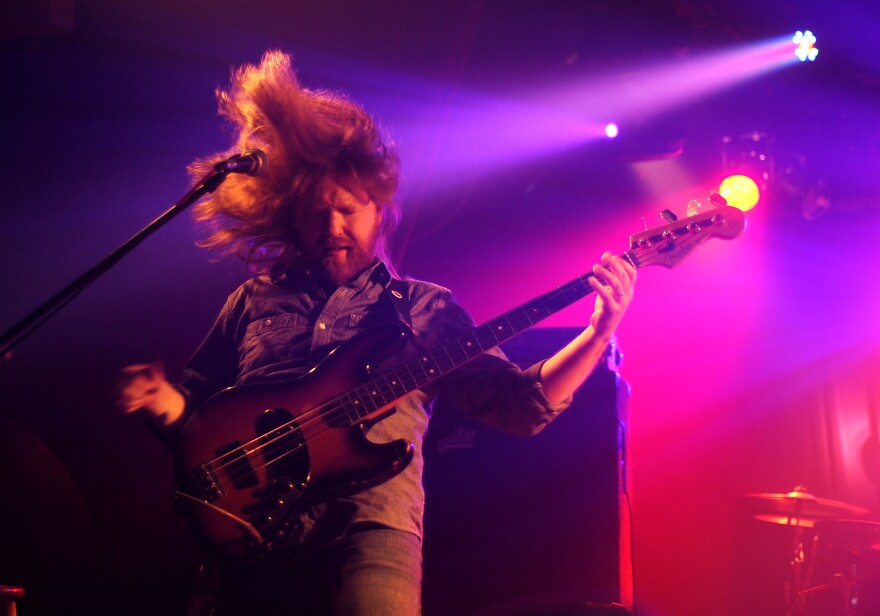 Bassist Reed Mathis flips his long hair on stage while playing guitar. He's under purple and red lighting.