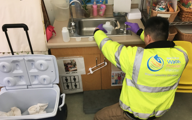 A person in a neon jacket and gloves kneels down and holds a bottle in a sink.