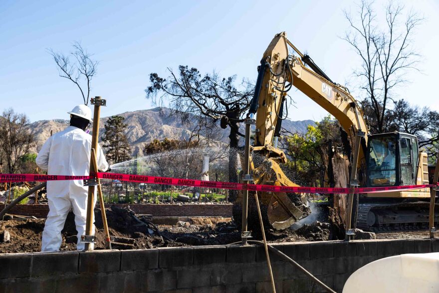 A man in a white coverall and helmet stands inside a debris area, with red danger tape surrounding the area. A backhoe clears ash and debris in the background.