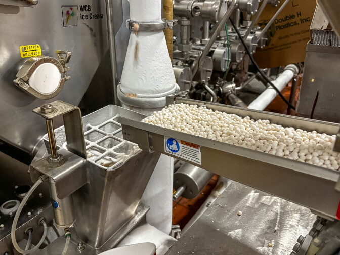 A metal feeder chute filled with small white malt balls, positioned above stainless steel ice cream production equipment.