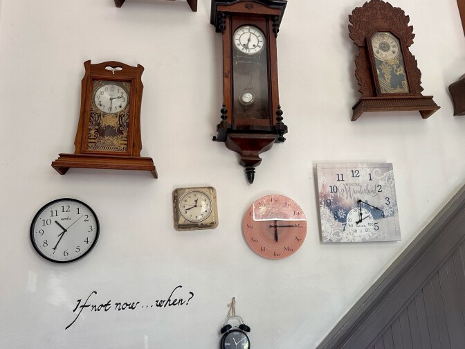 An array of clocks are on a white wall that also has the phrase "if not now...when?" written upon it.