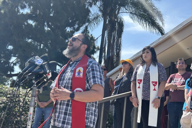A man with a beard wearing a red stole speaks into microphones at a news conference setting.