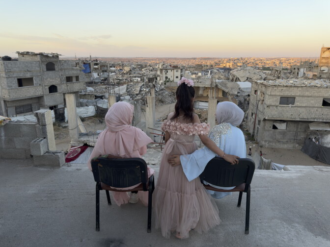 Three women are pictured from behind. Two are seated and one stands between them as they overlook a city of beige buildings at sunset.