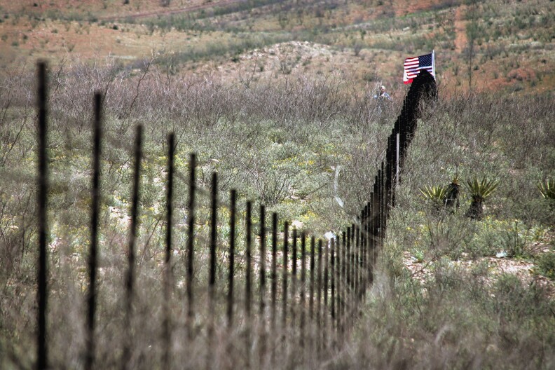 A volunteer from the Minuteman Project stands near an American flag placed in the barbed wire fence which divides the U.S./Mexican border April 4, 2005 near Noco, Sonora Mexico. More than a thousand volunteers from the Minuteman Project are expected to fan out across a 23 mile stretch of the Arizona border to search for Illegal aliens who are making the trek into the United States from Mexico during April. 