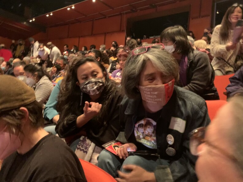 Two Asian American women, wearing masks, sit in a crowded theater. One of them leans forward to speak to a woman in another row.