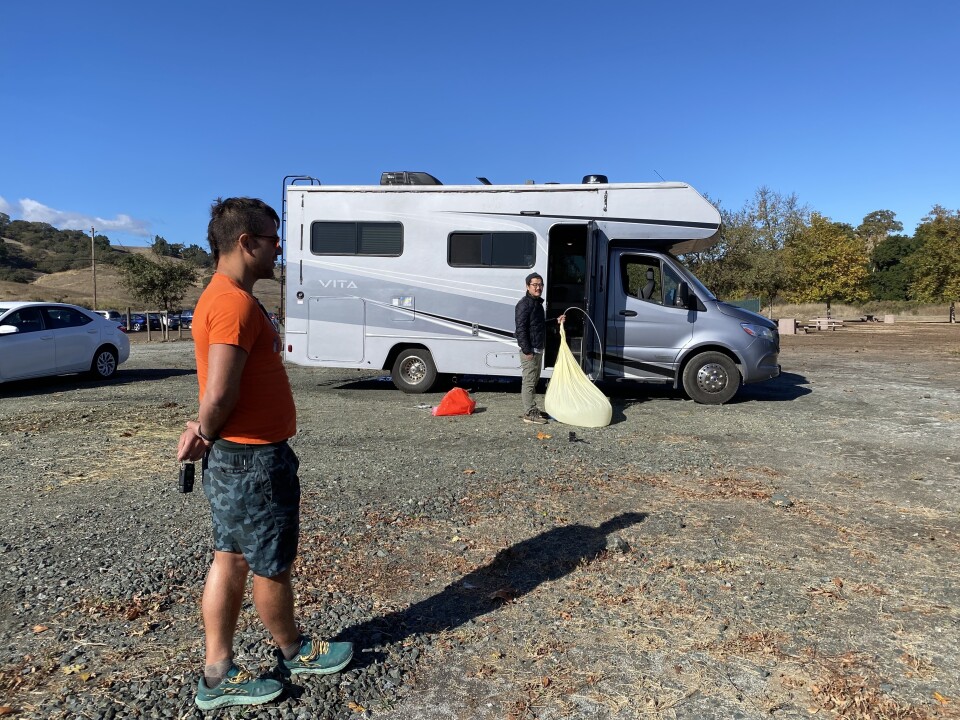 Two people stand near a silver camper van. One holds a deflated balloon.