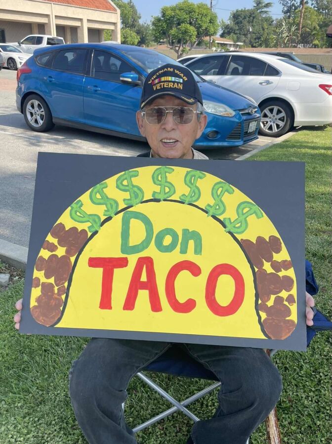 The image depicts a person seated outdoors on a chair, holding a sign that obscures their face. The person is wearing a hat labeled "VIETNAM WAR VETERAN." The sign they are holding features an illustration of a taco with dollar signs above it. The words "Don TACO" are written in green and red letters across the center. In the background, there are parked cars, suggesting the scene takes place in a public or urban setting.