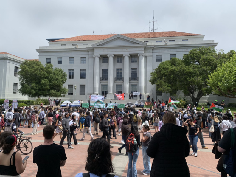 People cluster around an encampment in front of a mostly white building with columns.