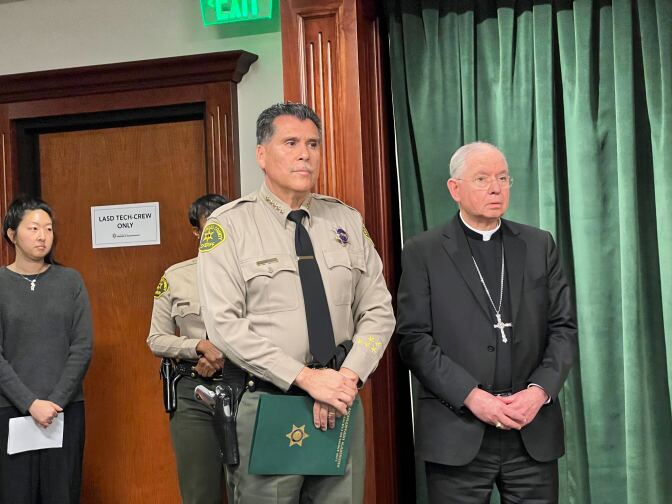 An older white man with a gray goatee and dressed in a Catholic bishop's white robe and vestment stands before an altar covered with a candles and a wooden crucifix. Parishioners stand over folding chairs in the background.