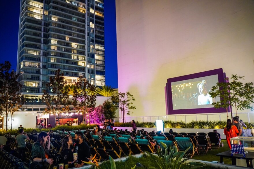 a handful of people sit in chairs on a high rise rooftop as a movie plays on a makeshift screen