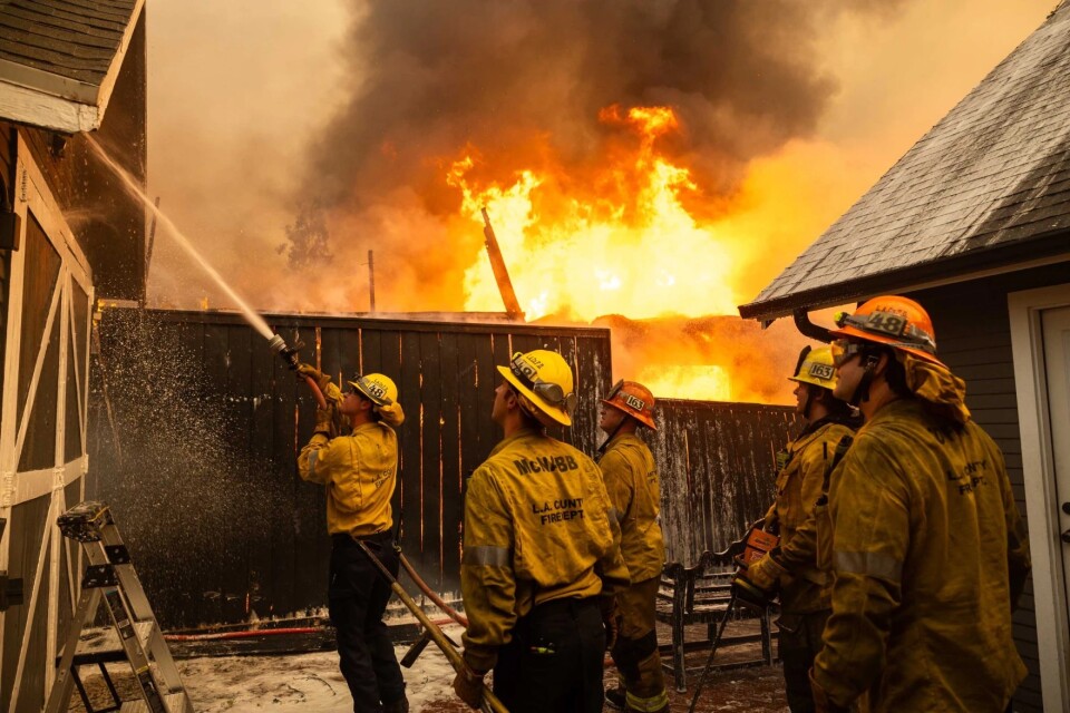 Firefighters spray water onto a home as a building while flames shoot high into the sky in the background, behind a wooden fence.
