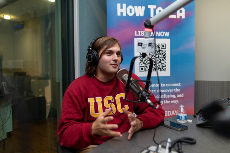 A masculine presenting person with light brown hair wearing a red sweatshirt with yellow lettering that reads "USC" speaks into a microphone in a recording studio.