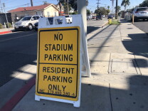 A yellow sign propped up with white plastic sits on the corner of a residential intersection in Inglewood. The sign reads: "NO STADIUM PARKING RESIDENT PARKING ONLY" in big letters, and in smaller letters at the bottom, says "IMC 3.79 AND 37.80" There are some cars parked on the street behind it. 