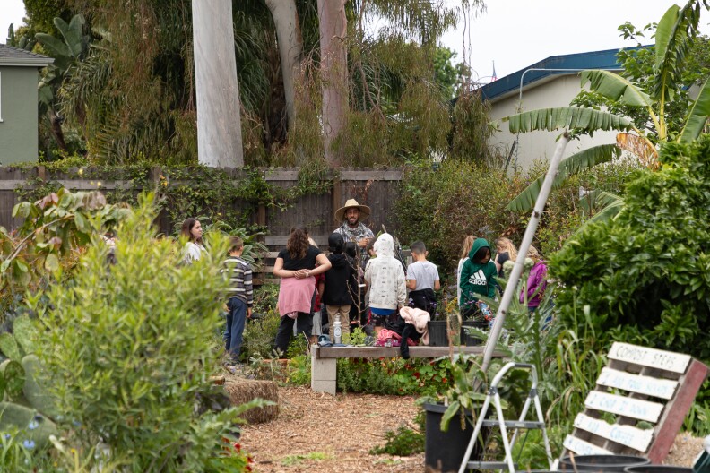 An outdoor garden where various young students stand facing their teacher.