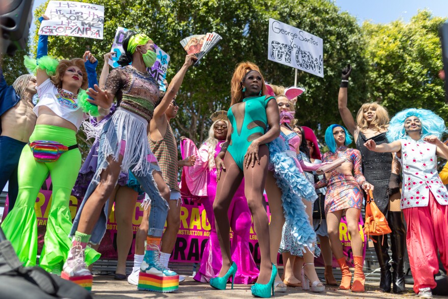 Multiple people in drag, one in a low plunge turquoise bathing suit and another wearing giant rainbow platform boots dance on a stage, holding placards protesting anti LGBTQ legislation.
