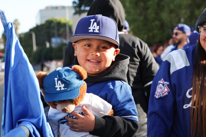 A little boy with medium-tone skin wearing a blue Dodgers hat and blue Dodgers jersey holds a teddy bear also wearing a Dodgers hat and jersey. 