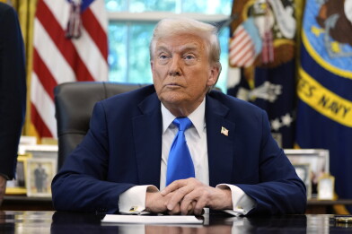 A man with a medium-light skin tone and white hair sits at a desk in a white shirt, a light blue tie and a blue blazer. He has an American flag pin on his lapel.