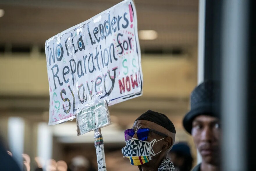 A Black man holds a sign that reads: "World Leaders! Reparations for Slavery NOW!" He is standing indoors, and another Black man stands next to him. The man holding the sign is wearing a black skull cap, sunglasses and a face mask with Black, white, yellow, red and green designs.