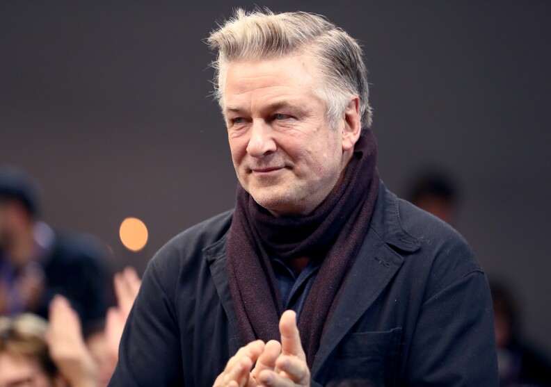 Actor Alec Baldwin, with white and gray hair, has a slight smile on his face. His hands are up in what looks like he's mid-applause. He wears a dark purplish scarf and a navy blue jacket, standing in front of others, some seated and some standing, at least one of whom appears to also be clapping.