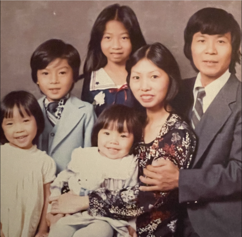 A Vietnamese family of six, with four children and a mother and a father pose for a family portrait. A young female with bangs and a bob smiles at the camera on the right, a young male in a light blue suit stands slightly behind her, slightly taller, and a young female with long hair and in a collared shirt stands in the center back. A female sits center-front, smiling as she holds a young infant with bangs. A male with dark hair in a gray suit stands behind the female, touching her arm.