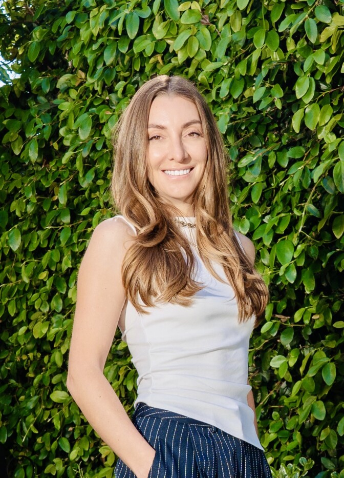 A woman with long hair stands against a wall of green foliage. 