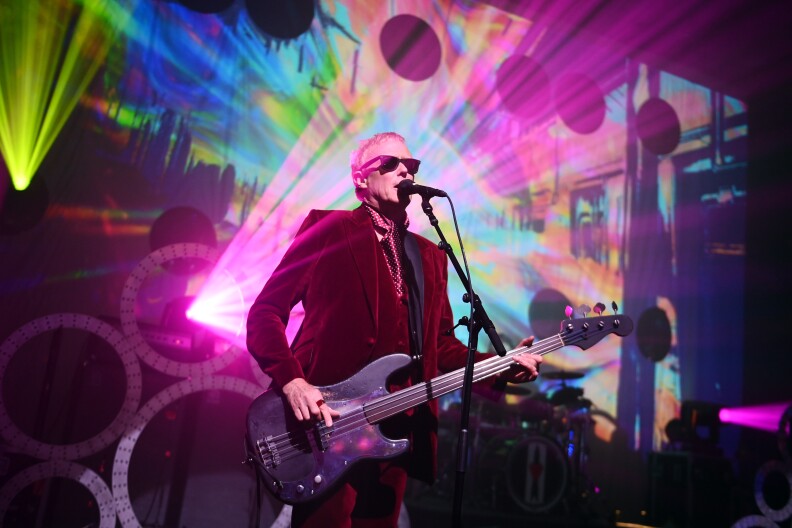 Image of artist David J on stage singing into a microphone and holding a guitar. Behind him is a dim lit stage while a bright pink light is coming out a machine, and also different colors on a back screen.
