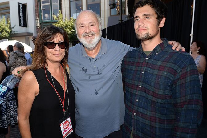 Director Rob Reiner, a man with light skin tone, bald head and white beard, smiles as he stands in between and hugs his wife, Michele Singer, a woman with light skin tone, wearing a black dress and sunglasses, and son, Nick Reiner, a man with light skin tone, short goatee, wearing a dark-colored flannel. They pose for a photograph with Rob Reiner and Michele Singer look at the camera, while Nick Reiner looks away.