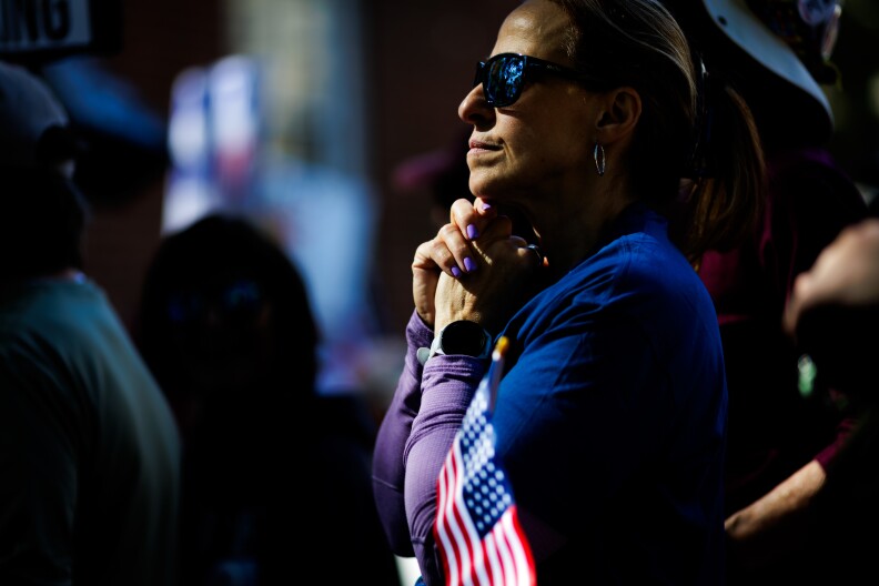A woman clasps her hands together. A U.S. flag is in the crook of her arm.