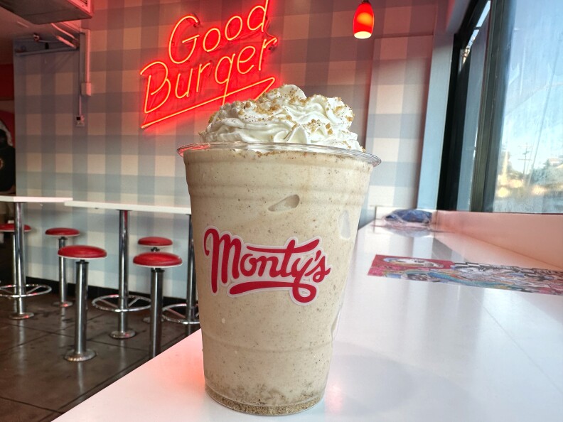 A milkshake in a clear plastic cup with a red-lettered logo and the name "Monty's" sits on a white countertop next to a window. In the background, there are a group of white high-topped tables with metal stools featuring circular red vinyl cushions. Above them, there is a large red neon sign that hangs on the wall, reading "Good Burger." The wall has light blue and white wallpaper.