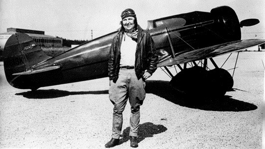 A black and white photo of a fair-skinned women standing in aviator uniform posing in front of an airplane.