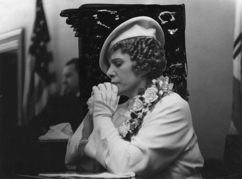 Aimee Semple McPherson, her eyes closed and hands folded, seated in what appears to be a courtroom. Circa 1937.