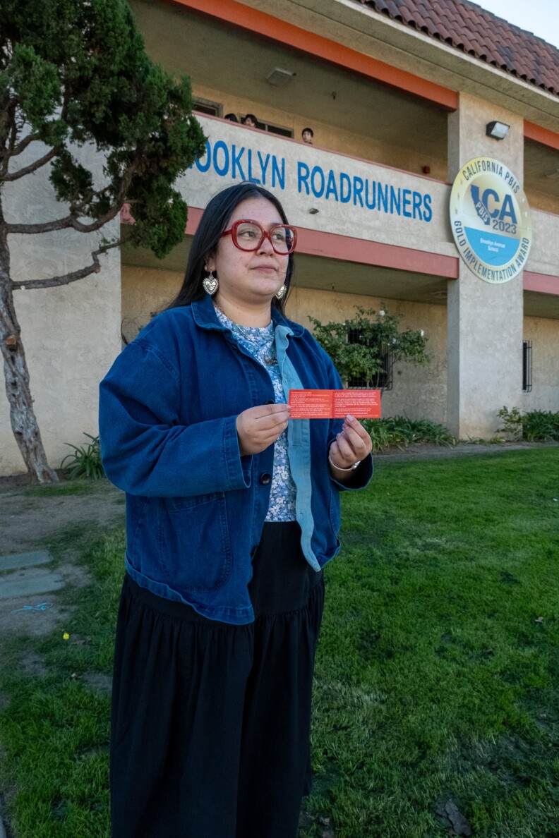 A female presenting person hearing a denim shirt and red glasses holds two red cards next to each other. She stands on a lawn in front of a building with text that reads "BROOKLYN ROADRUNNERS" and an emblem next to that.