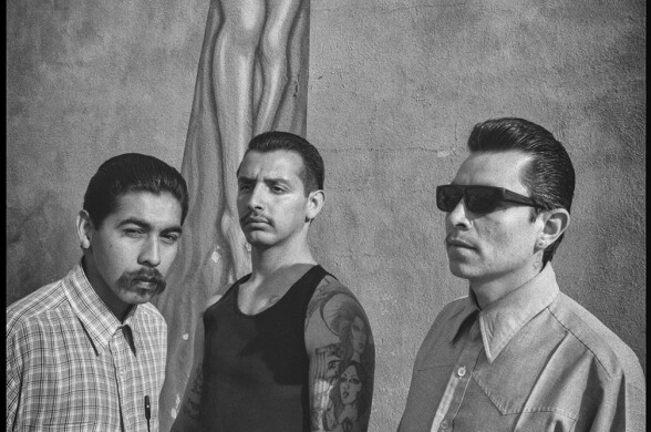 Three Latino men with short dark hair stand in front of a mural of a crucifix in daylight. The photo is black and white.