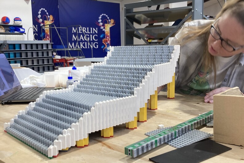 A woman with long hair works on Lego bleachers, mostly in gray and white, on a work table. Behind her a blue banner reads MERLIN MAGIC MAKING.
