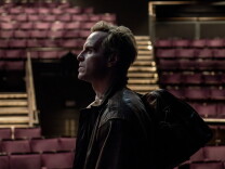 Side profile view of Andrew Scott standing in an auditorium. Red seats can be seen in the background.