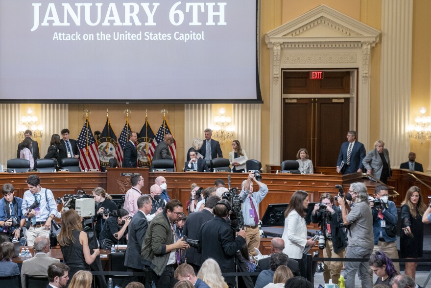 Photographers take photos of a woman walking away from a hearing room as a large screen displays the words January 6th in the background.