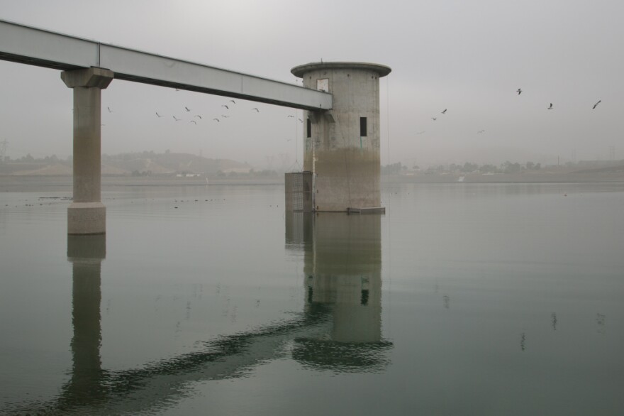 A gray cylindrical structure rises from the middle of a reservoir on a gray, gloomy day. A raised aqueduct leads directly into the side of the structure.
