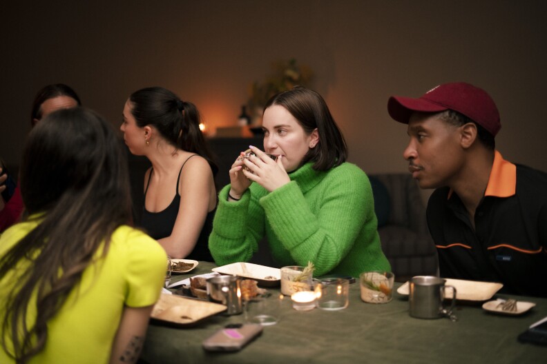 A light-skinned woman sits with her elbows on a table, wearing a bright green turtleneck sweater. She's holding a cup with both hands up to her face, about to take a drink. A man with dark skin sits next to her and is wearing a red baseball cap and a black shirt with an orange collar.