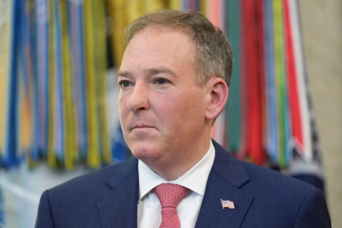 Man with light-toned skin, short brown and gray hair, looking off to the side. He is wearing a dark navy suit jacket over a white collared shirt, a red tie with a small repeating pattern, and a small American flag pin on the lapel.