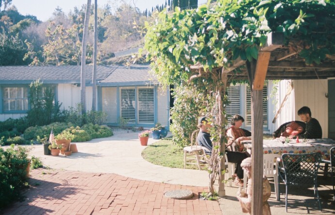 A family sits outside under an awning covered in green vines. It's a sunny day. A house and palm trees are in the background.