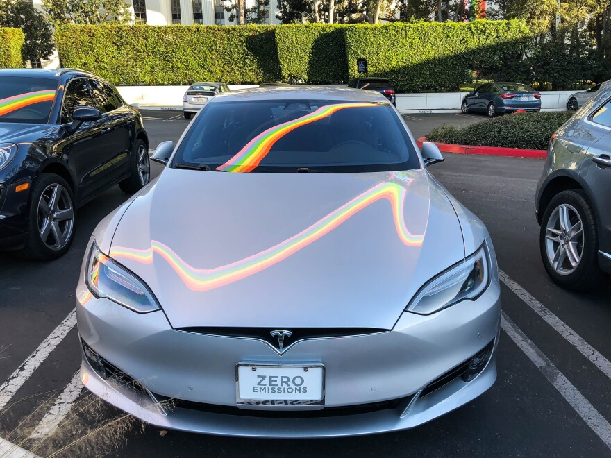 A silver Tesla electric vehicle has a colorful reflection on it.