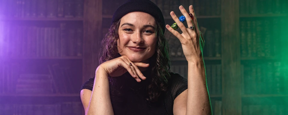 A light-skinned woman wearing black smiles and holds 3 20-sided dice between her fingers. 