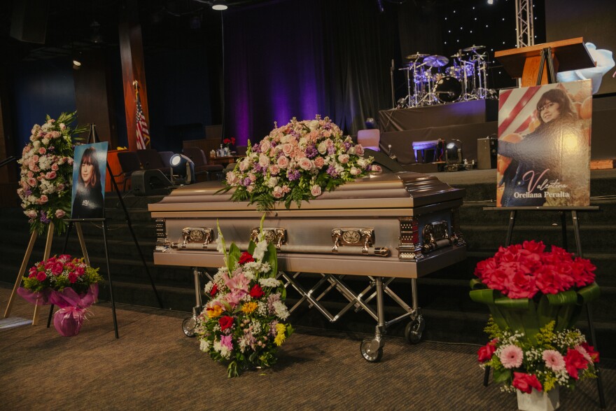 Large flower arrangements sit on and near Valentina Orellana Peralta's coffin. A large photographs of her stands on easels on either side of the coffin. A drum set is on a stage in the distant background.