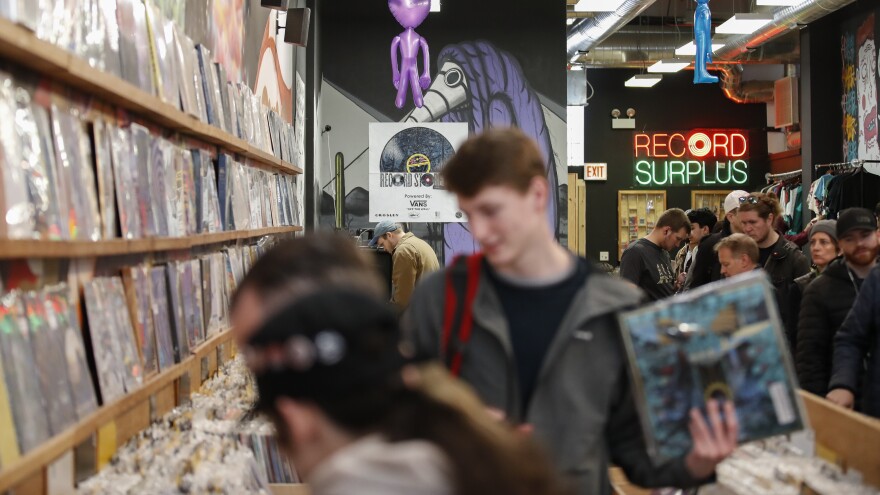 Three customers are shown blurred as they look through records at a record store. A large crowd of people can be seen behind them as well. A neon sign reads "Record Surplus" in red and green lights.