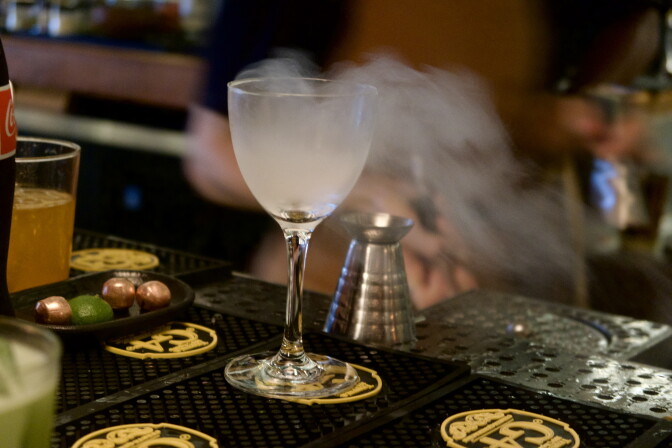 Smoke swirls above a stemmed cocktail glass on a bar mat, next to a metal jigger and a dish of limes.