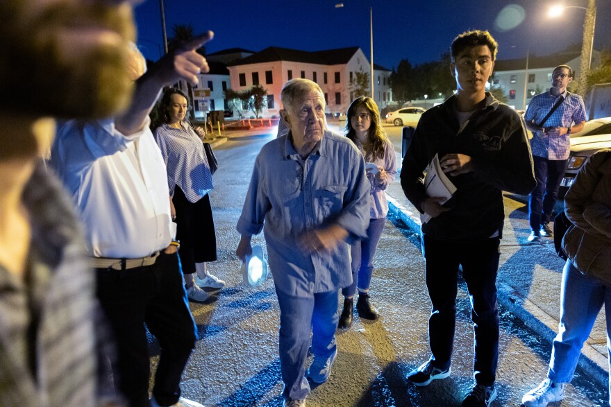 A man walks at night in a blue outfit surrounded by other people, along a road with buildings in the background, as someone points in the direction the man is looking.