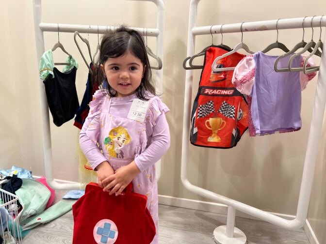 A female with light skin tone and dark hair wearing a purple dress and holding a red bag looks ahead. She is standing in front of a clothing rack with different costumes. 