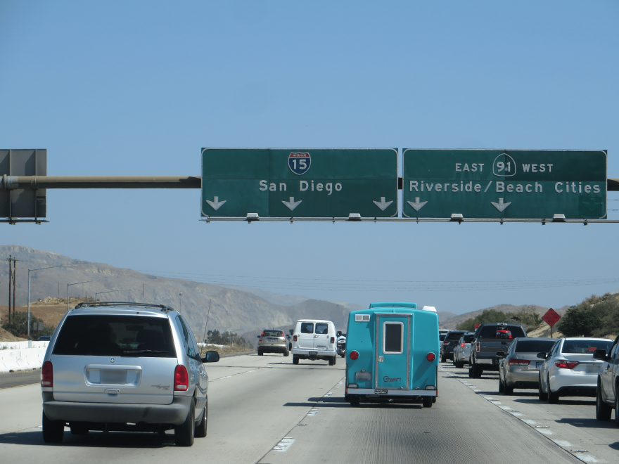 Cars travel on a gray four lane freeway below to overhead road signs that read "15 San Diego" and "East and West 91 Riverside/Beach Cities" to the left of a tan mountain range in front of a blue sky.